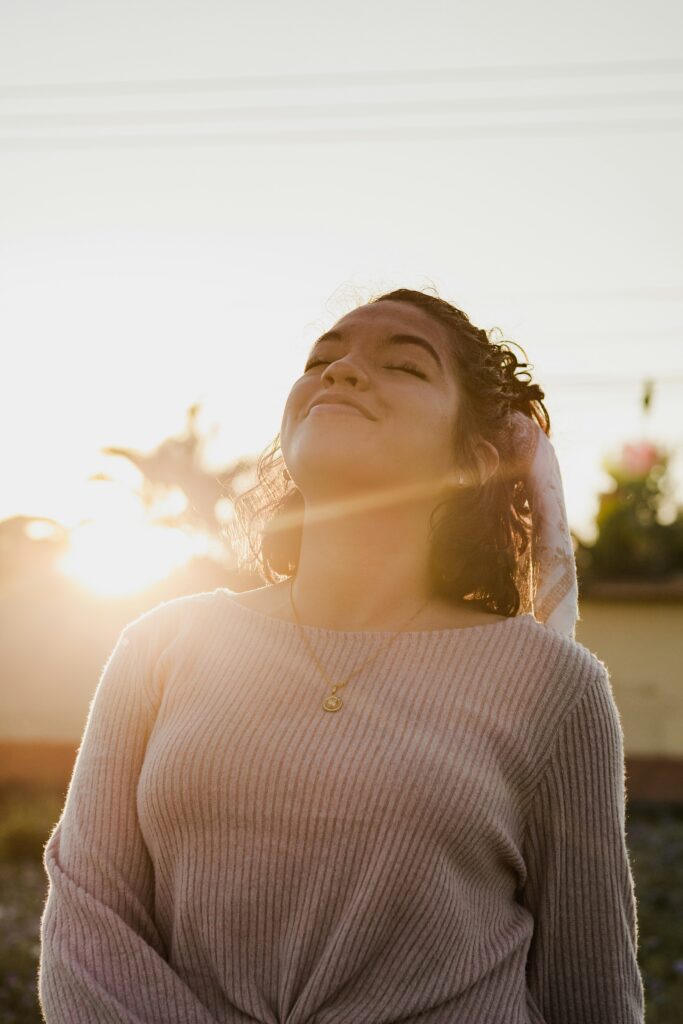 Mulher jovem de olhos fechados sorrindo ao pôr do sol, simbolizando amor próprio, autoestima feminina e o ato de se priorizar sem culpa